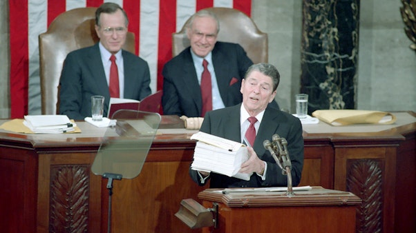 President Reagan speaks about Domestic Policy President Reagan speaking at a podium with two men sitting behind him at a desk in front of the American flag