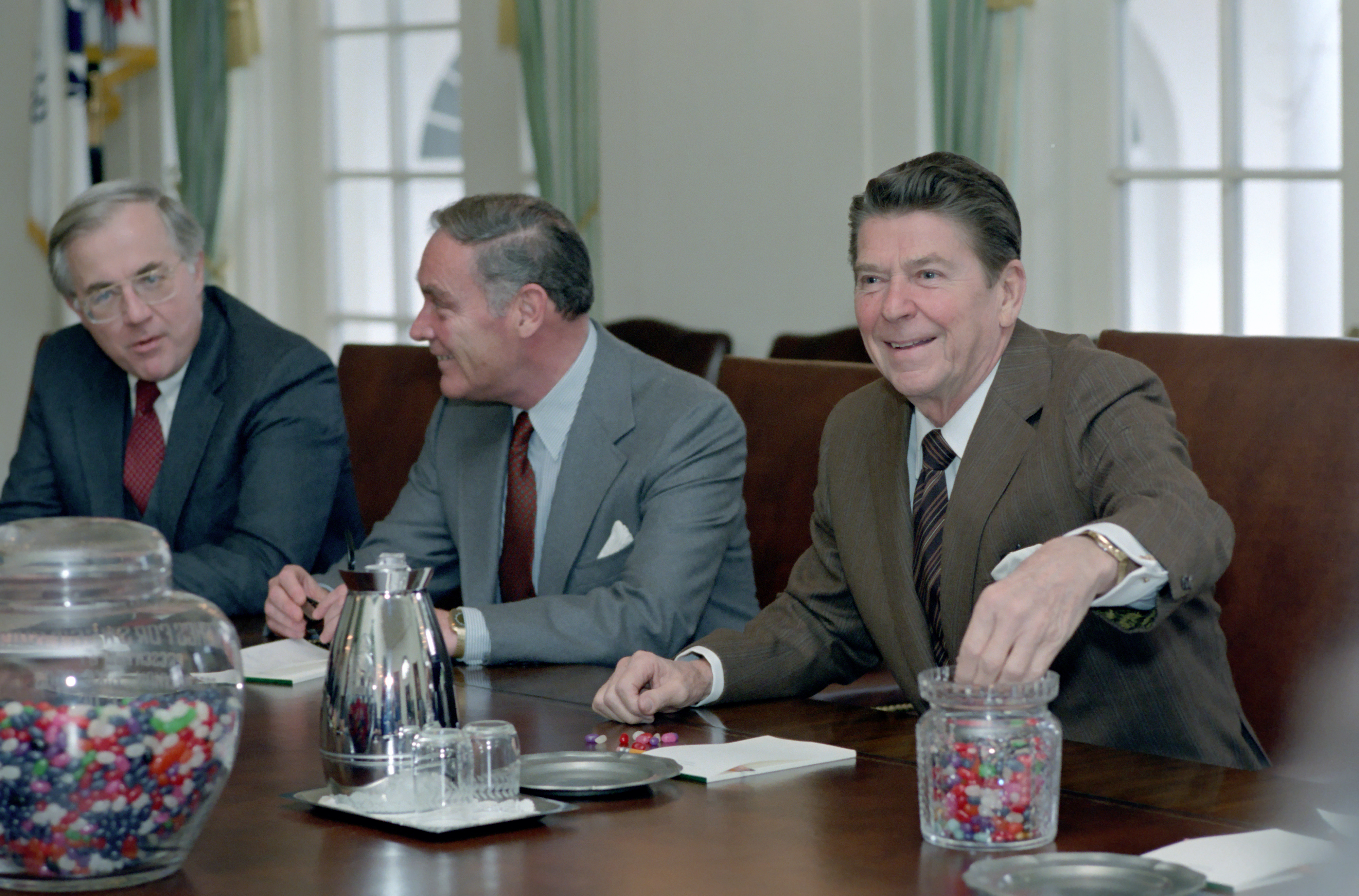 Giant jar of jelly beans at a table of three men with Ronald Reagan reaching into a smaller jar of jelly beans