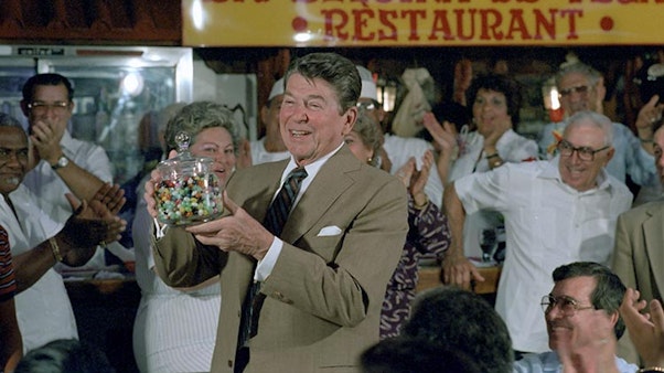 President Reagan receiving a jar of jellybeans during a Lunch at La Esquina de Tejas Restaurant in Miami, Florida. 05/20/1983. President Reagan in a tan suit holding a jar of jelly beans in front of a crowd of people