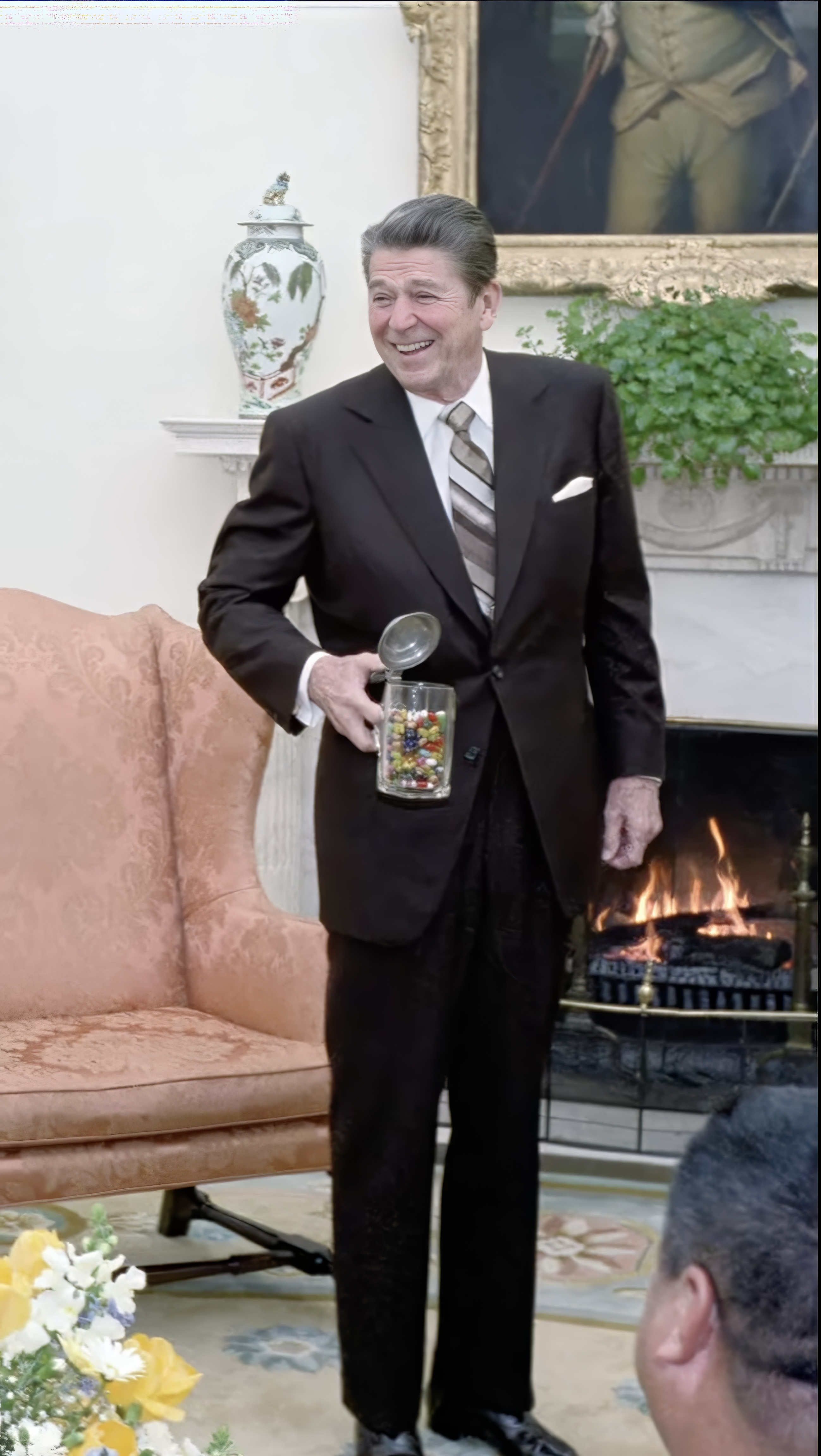 President Ronald Reagan smiling holding a jelly bean jar in the Oval Office in front of a fireplace