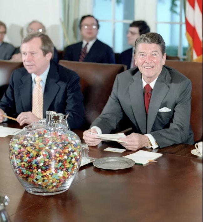 Reagan in meeting with giant jelly bean jar Photo of President Reagan sitting with people at a large brown table with a giant colorful jelly bean jar